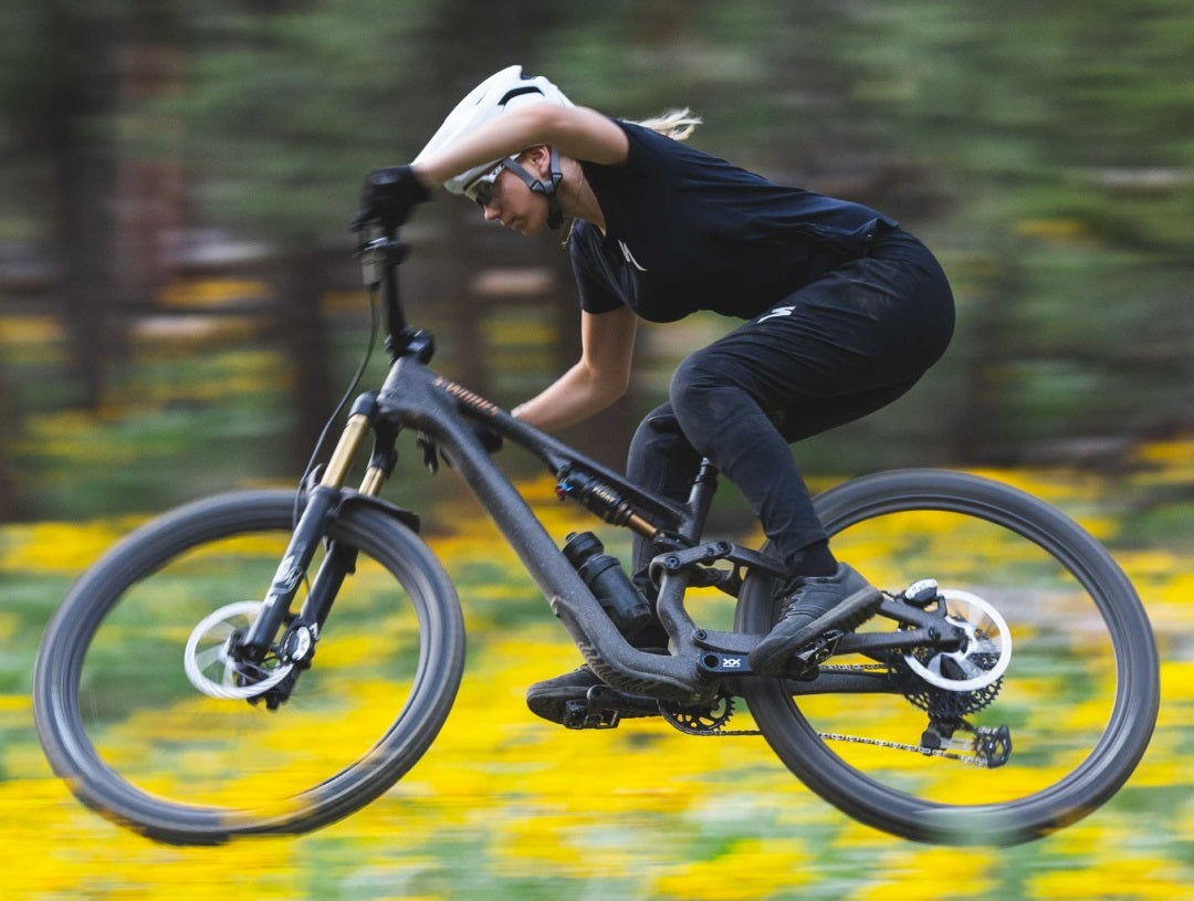 Person springt auf einem Specialized Stumpjumper Mountainbike in schwarzer Kleidung und Helm durch eine blühende Wiese.