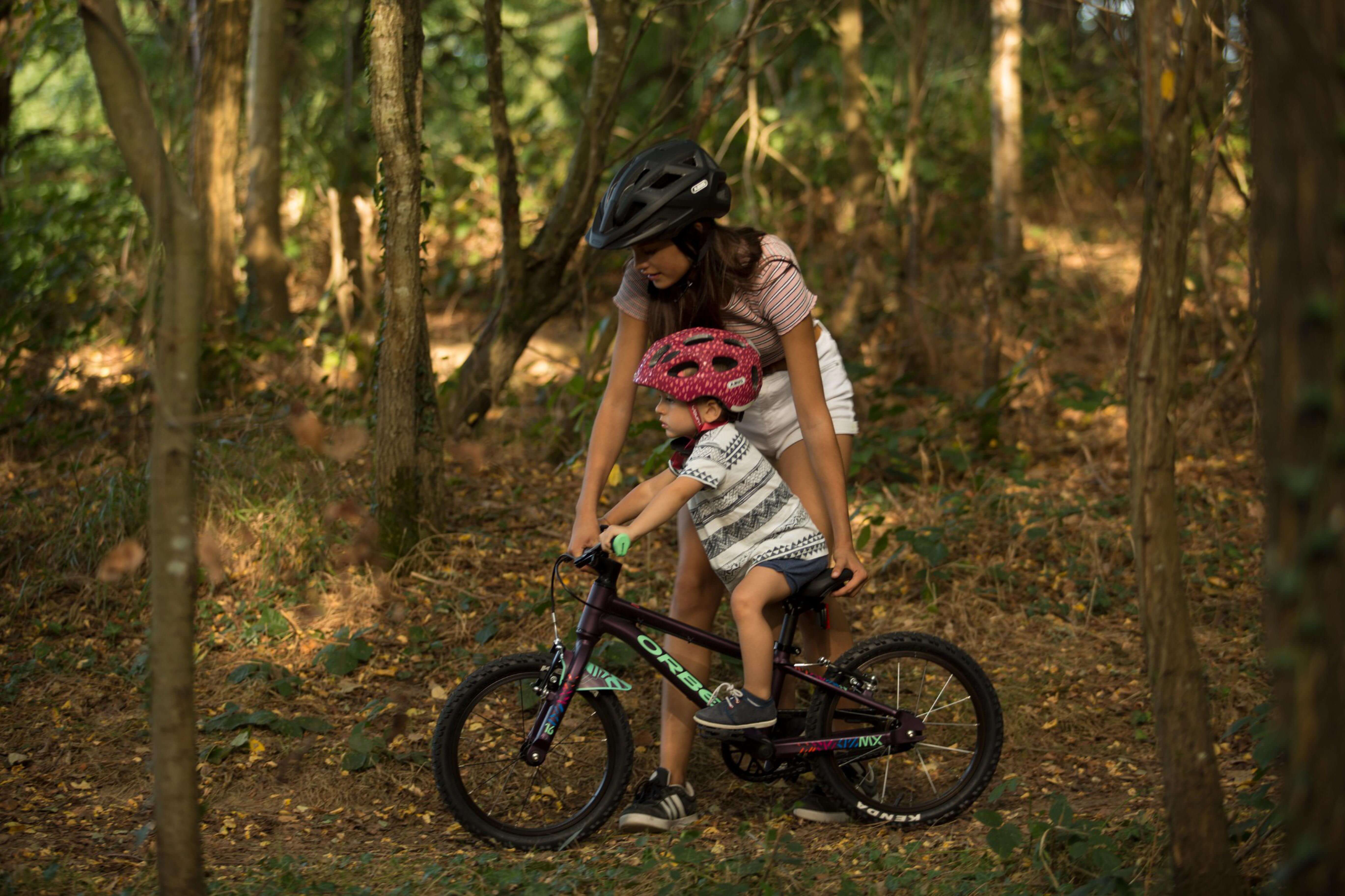 Kind mit rosa Helm fährt unter Aufsicht ein Orbea MX Junior Fahrrad im Wald.
