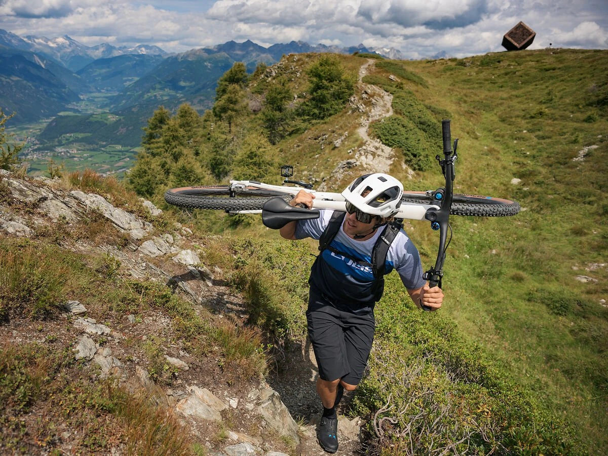 Person trägt ein Mountainbike auf einem Bergpfad; malerische Landschaft mit Bergen und bewölktem Himmel im Hintergrund.