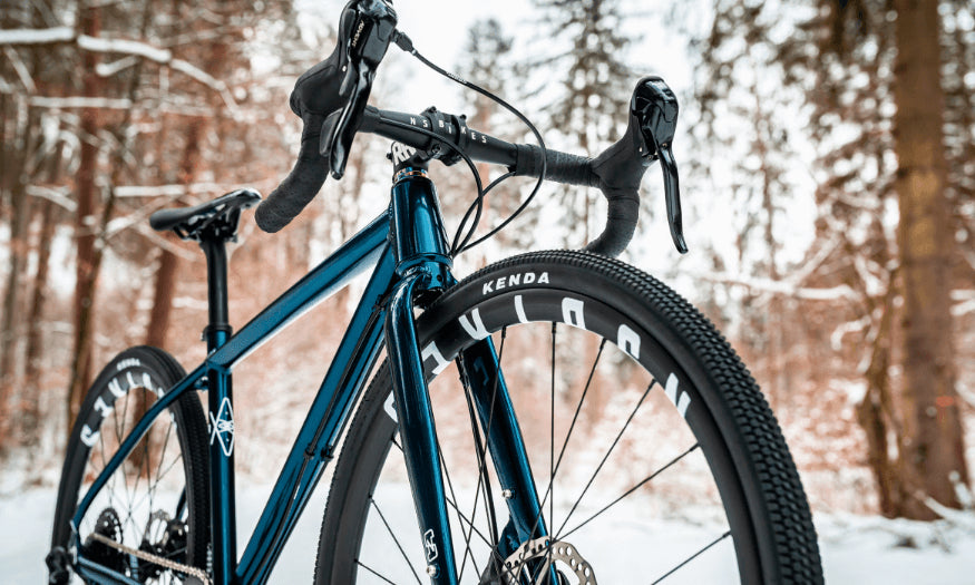 Gravelbike auf verschneitem Waldweg, blaue Rahmenlackierung, dicke Kenda-Reifen, geeignet für Offroad-Abenteuer.
