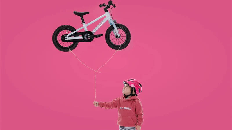 Kind mit rosa Helm, hält ein schwebendes, weißes Kinderfahrrad von Cube vor pinkem Hintergrund.
