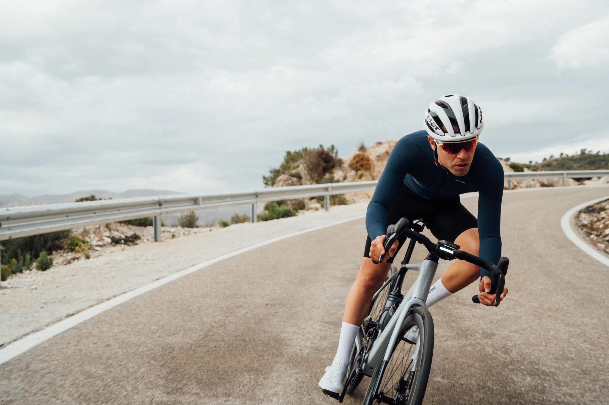 Radfahrer mit Helm auf einem Orbea Gain Rennrad auf kurviger Bergstraße bei bewölktem Himmel.