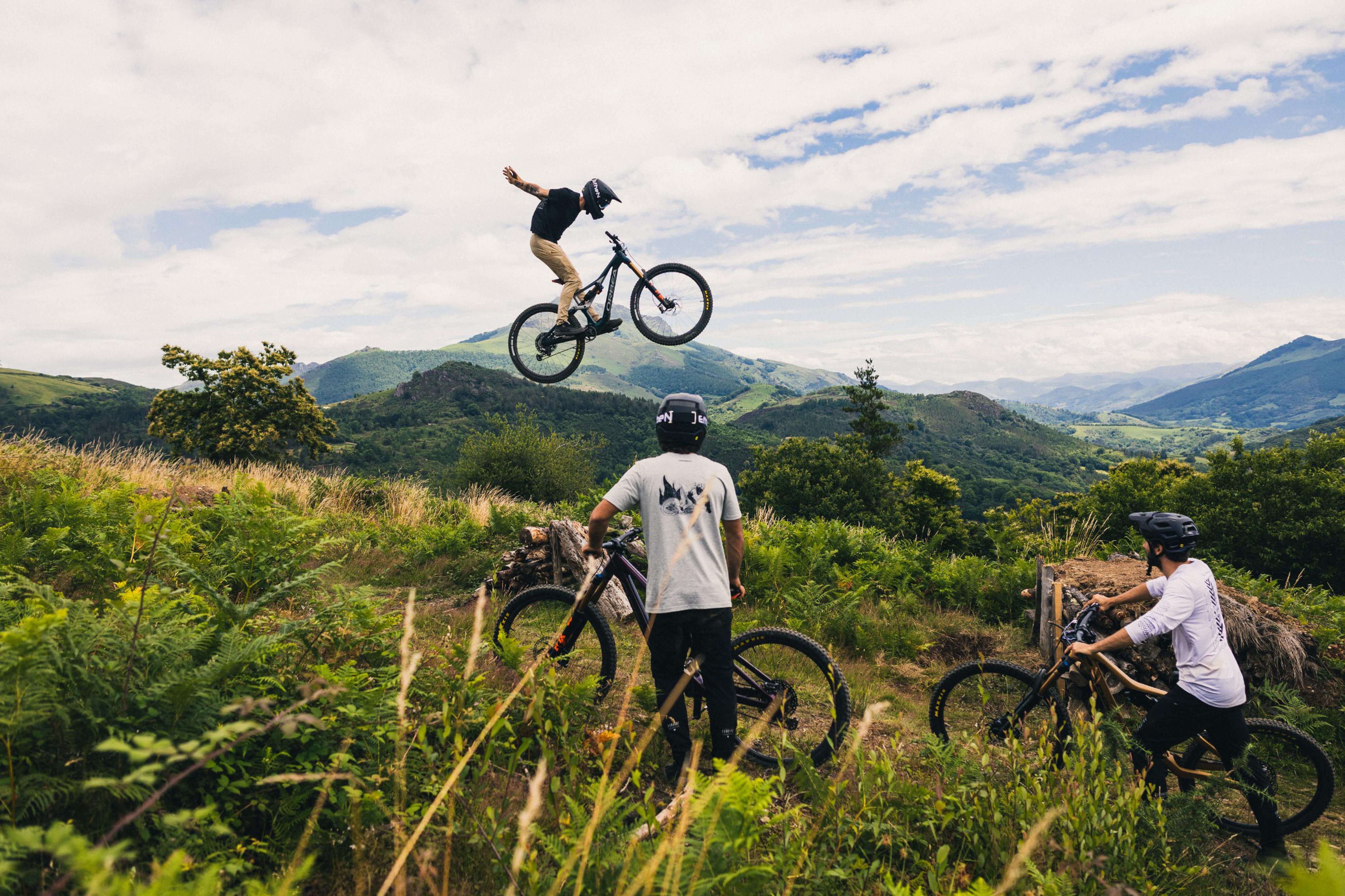 Mountainbiker springt mit Orbea Fully über Hügel, Zuschauer mit Helmen in schöner Landschaft. Perfekt für Abenteuersport.