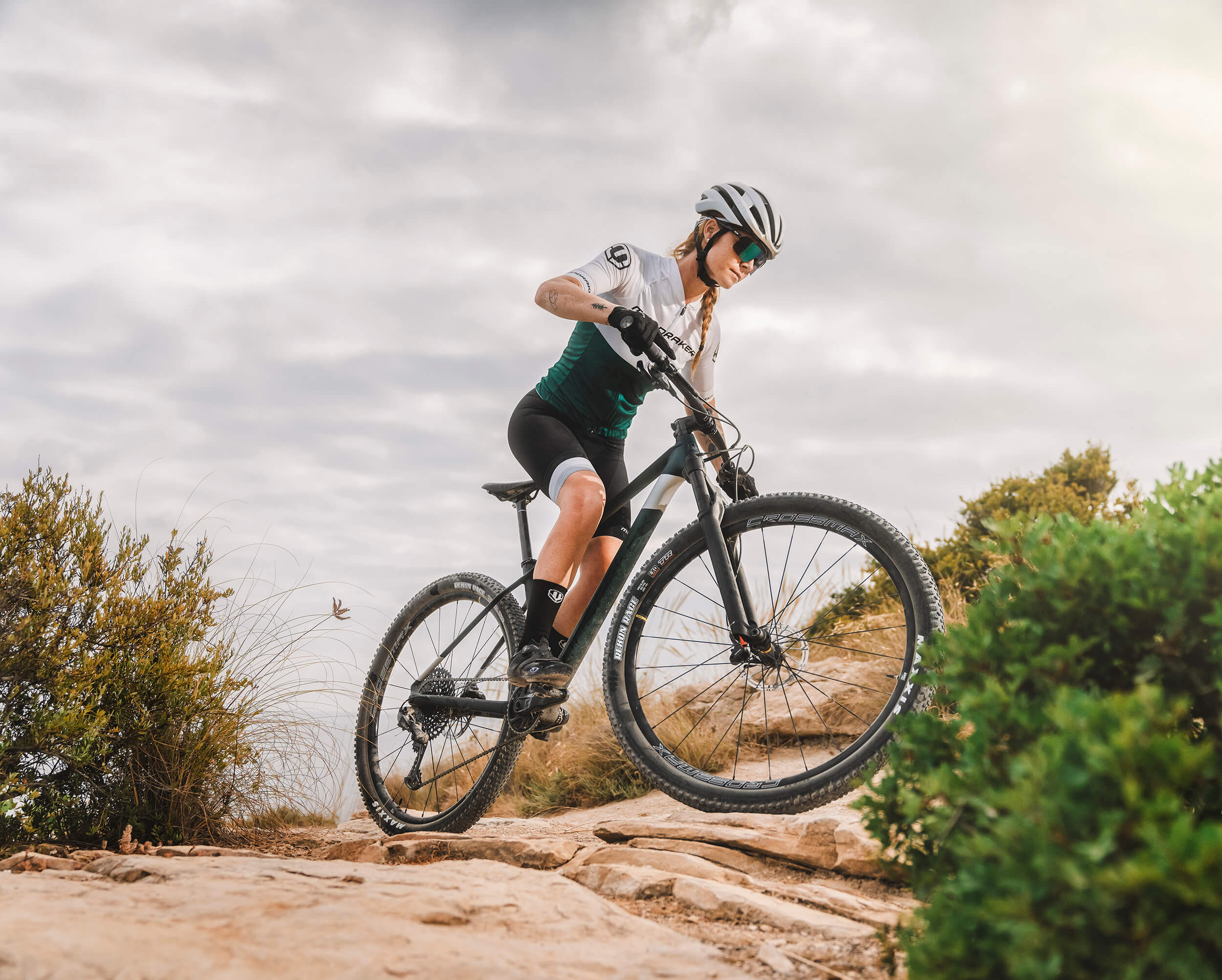 Bikerin mit Helm auf Mondraker Mountainbike fährt über felsigen Pfad in natürlicher Landschaft unter bewölktem Himmel.