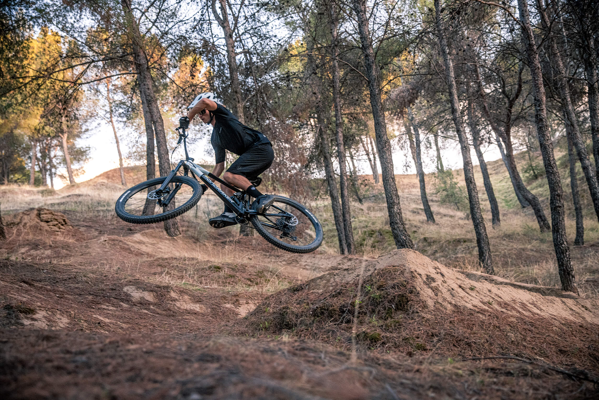 Person springt auf Mondraker-Mountainbike im Wald, trägt schwarzen Helm und Kleidung, geeignet für Mountainbike-Enthusiasten.