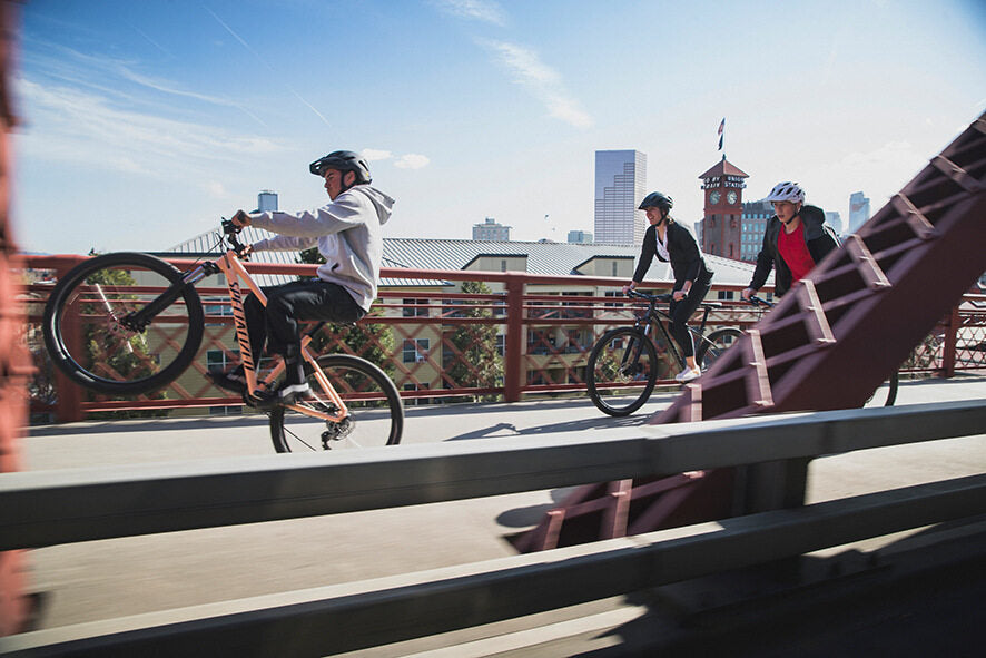 Drei Personen fahren auf Specialized Mountainbikes über eine Brücke in der Stadt; eine Person macht einen Wheelie.