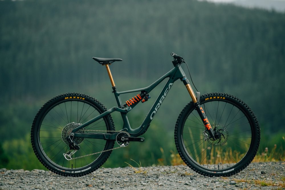 Mountainbike Orbea im Profil, dunkelgrün, mit Maxxis-Reifen, geeignet für Offroad-Abenteuer in bergiger Umgebung.