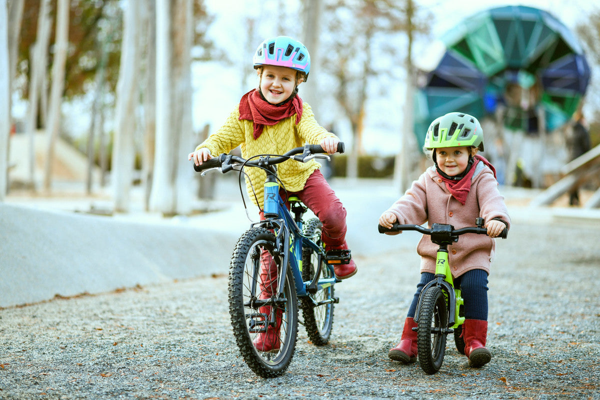 Zwei Kinder mit Helmen fahren Fahrräder und Laufrad auf einem Parkweg, lächelnd und warm gekleidet.