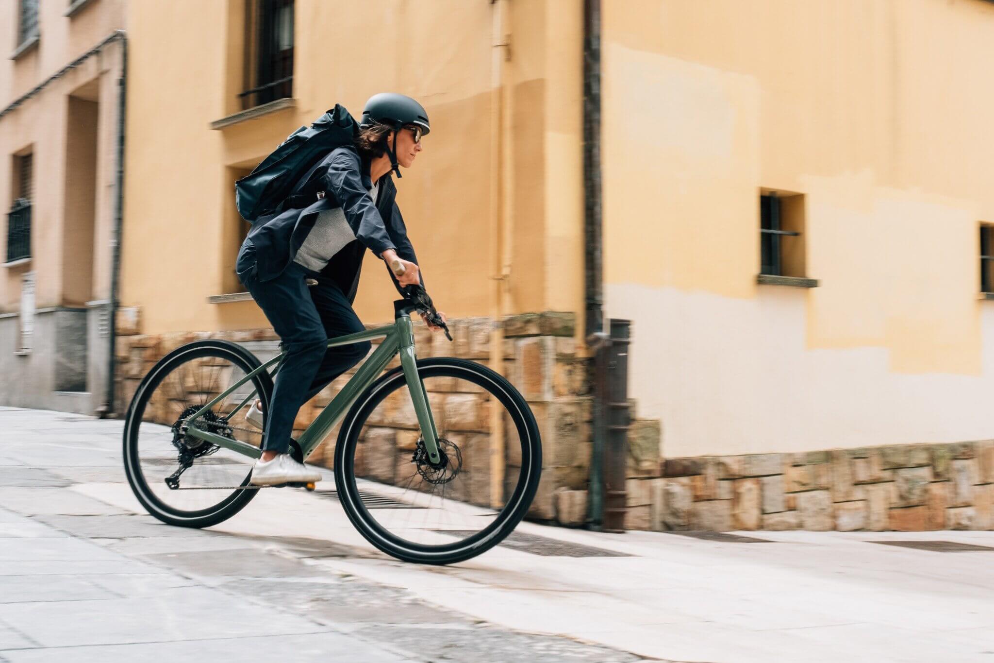 Person fährt ein Orbea Vibe Fahrrad in urbaner Umgebung, ideal für Pendler. Trägt Helm und Rucksack.
