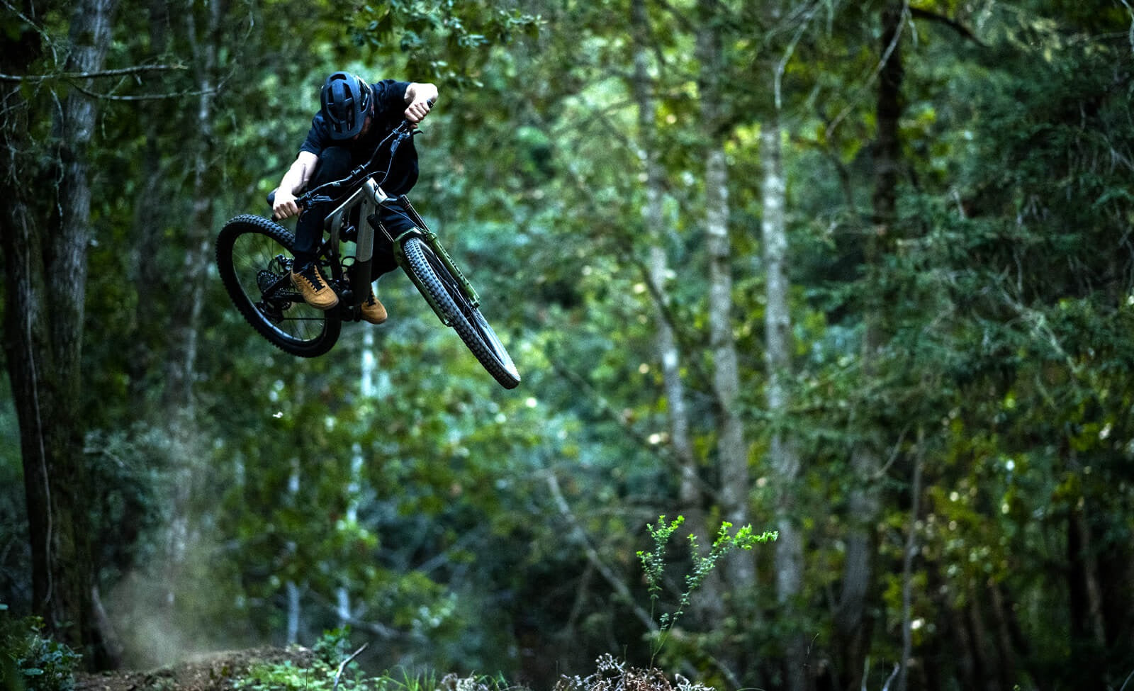 Mountainbiker in der Luft springt durch Waldgebiet mit full-suspension Fahrrad; trägt Helm und Schutzkleidung.