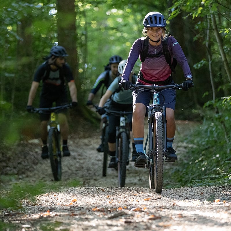 Frau auf Cube Access WS Mountainbike im Wald, gefolgt von drei Personen auf Fahrradtour, sportliche Kleidung und Helme.