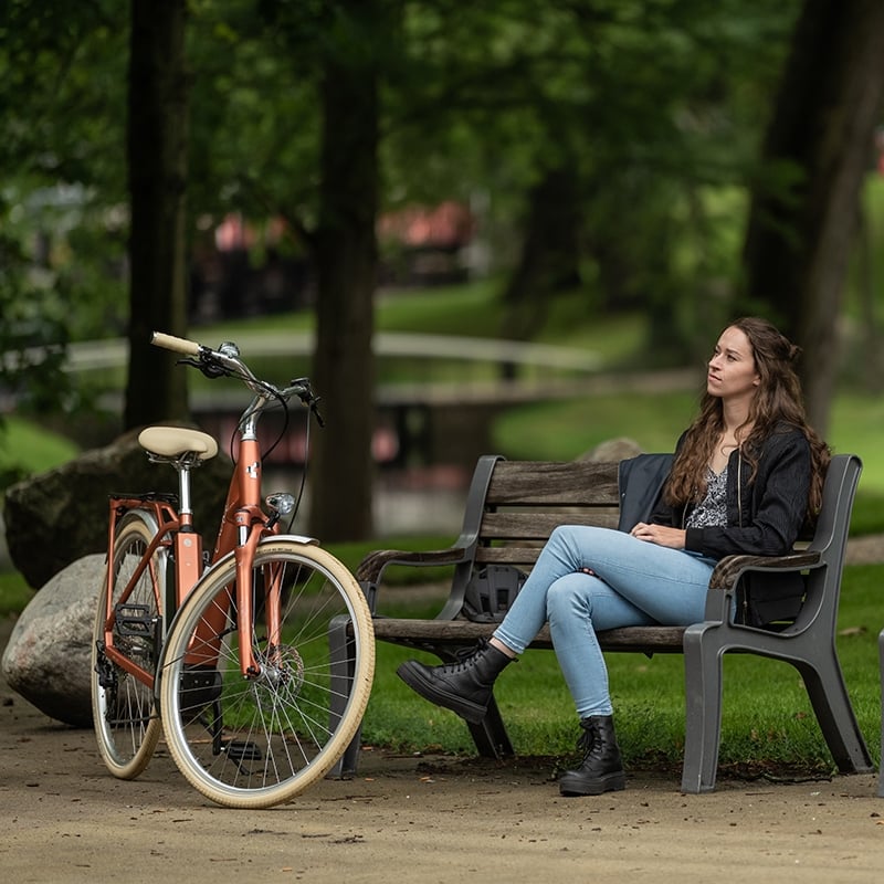 Frau auf Parkbank neben einem orangefarbenen Stadtfahrrad, geeignet für urbane Mobilität.