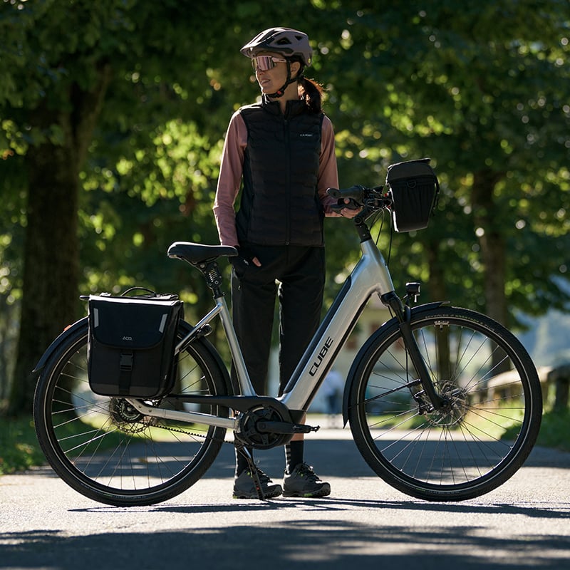 Frau mit Helm steht neben CUBE Supreme Hybrid E-Bike auf einem Weg, geeignet für Stadtfahrten, mit Taschen ausgestattet.