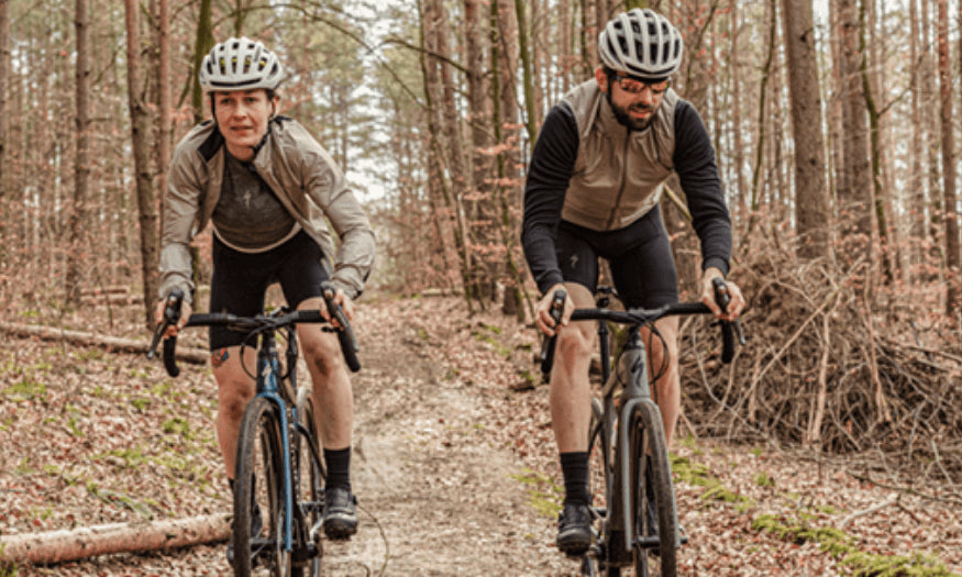 Zwei Radfahrer auf Gravelbikes im Wald. Beide tragen Helme und sportliche Bekleidung, geeignet für Offroad-Strecken.
