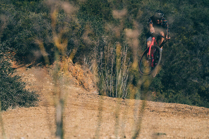 Radfahrer auf Specialized Gravelbike springt über Schotterpiste in gebirgiger Landschaft, ideal für Outdoor-Enthusiasten.