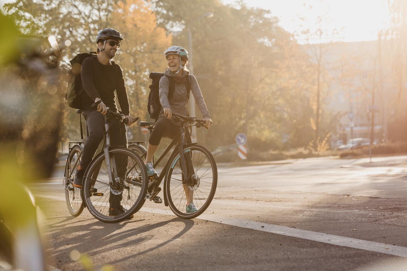 Zwei Radfahrer auf Stadtfahrrädern mit Helmen und Rucksäcken bei sonnigem Wetter.