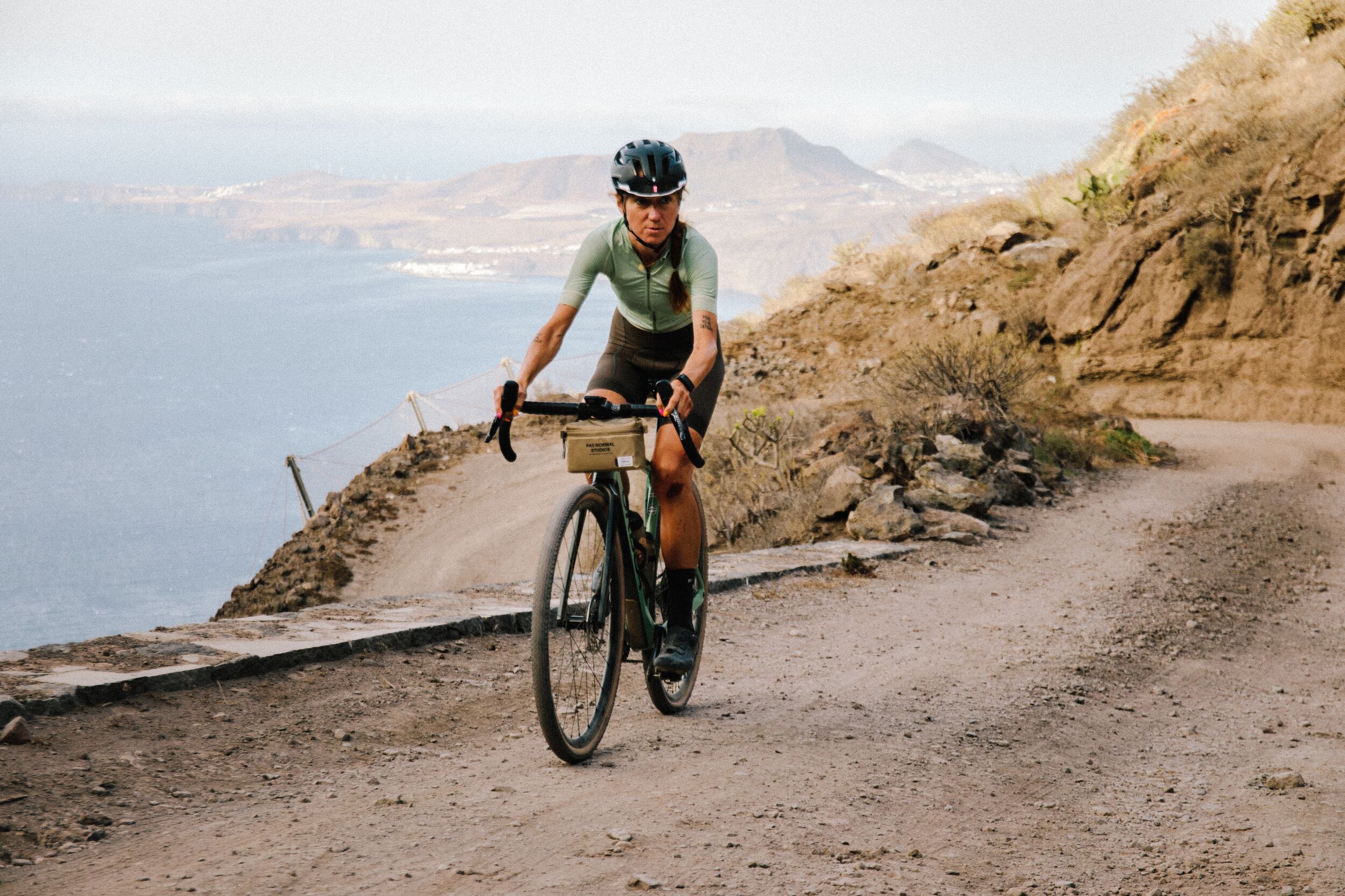 Fahrradfahrer auf einem Schotterweg mit Blick auf das Meer, Gravelbike, bergige Landschaft im Hintergrund.