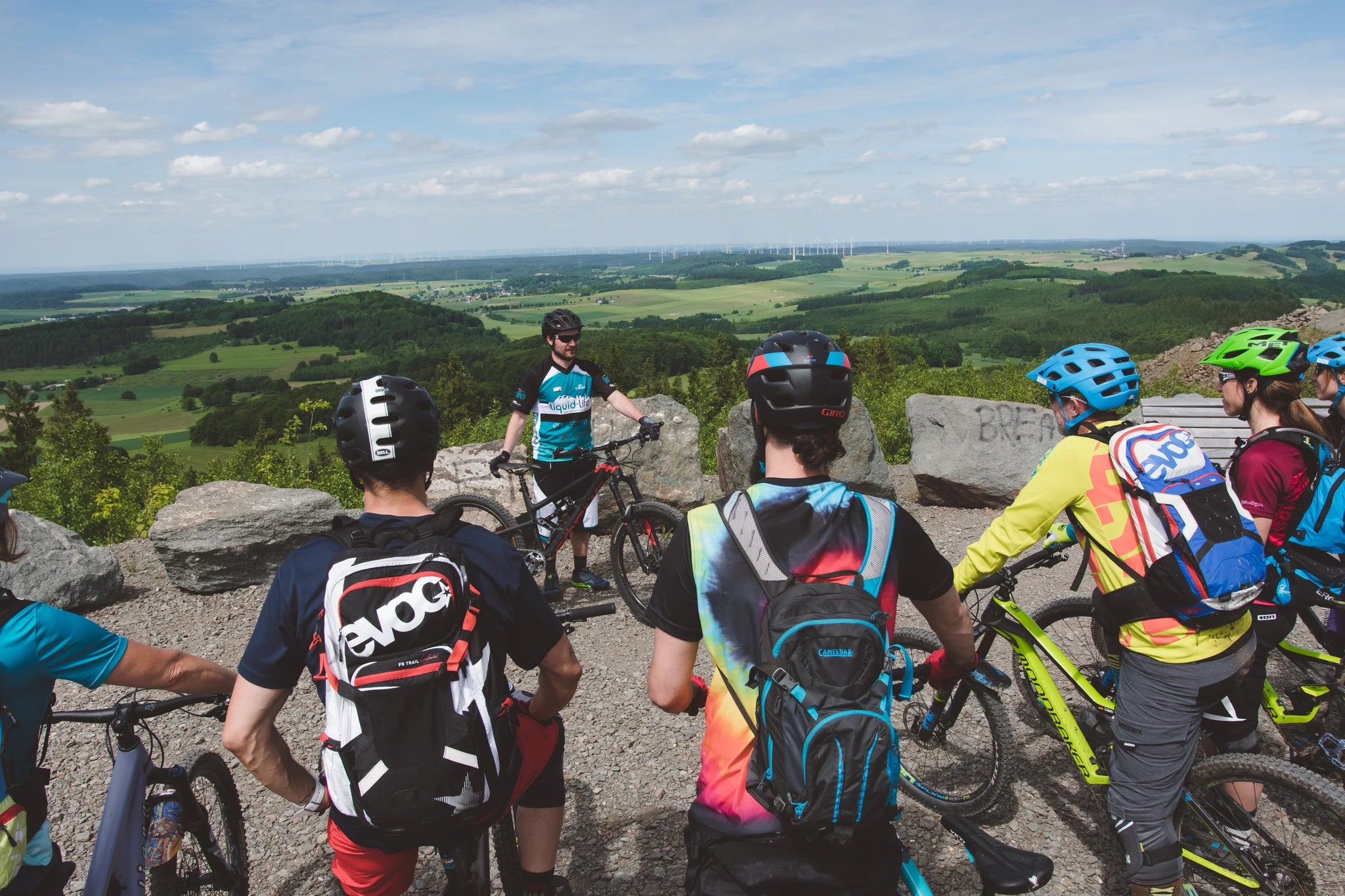 Gruppe von Mountainbikern mit Helmen und Rucksäcken bei einer Rast auf einem Aussichtspunkt mit weitem Blick auf die Landschaft.
