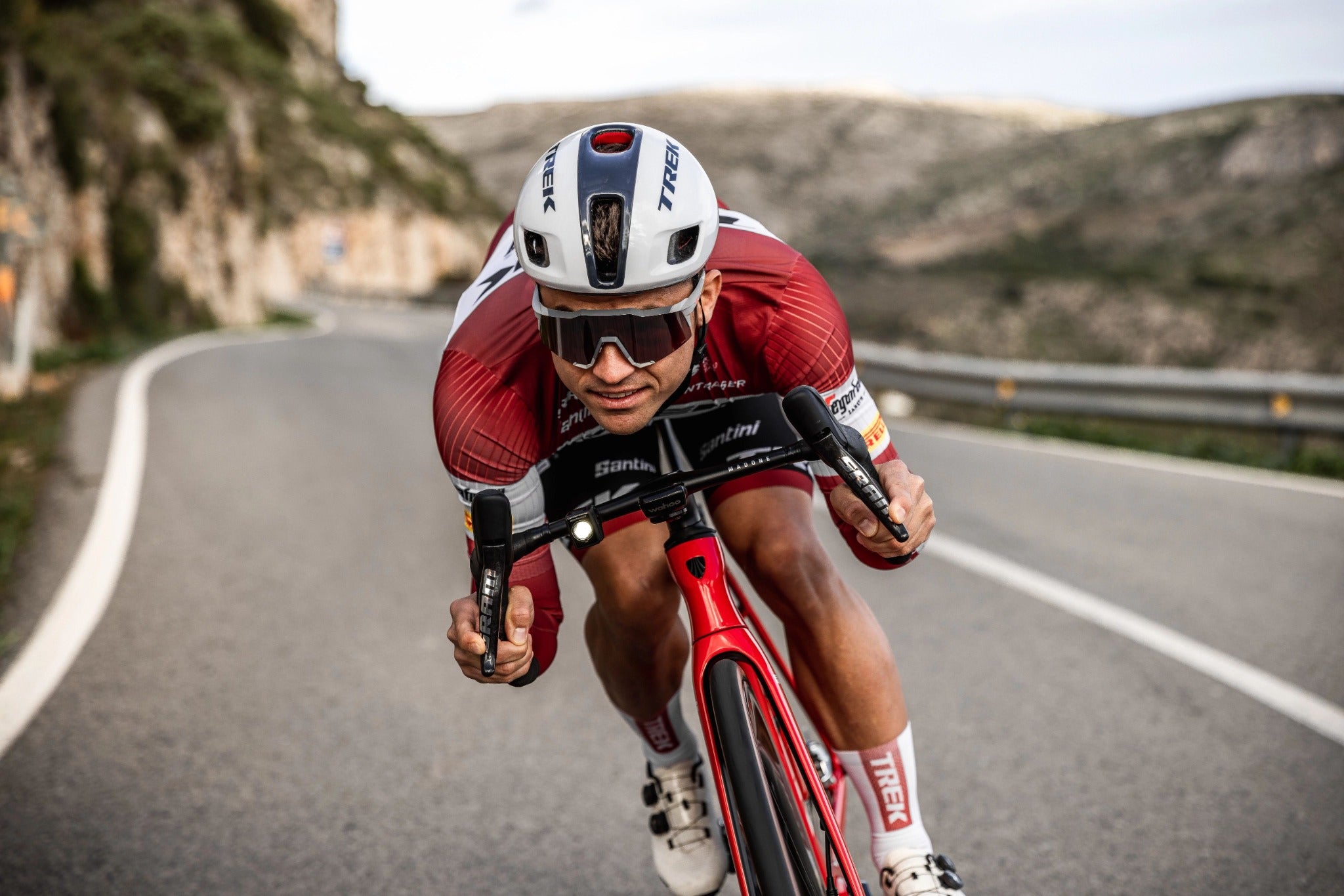 Radfahrer in Rennbekleidung auf einem Trek-Rennrad, fährt auf kurviger Straße durch hügelige Landschaft.