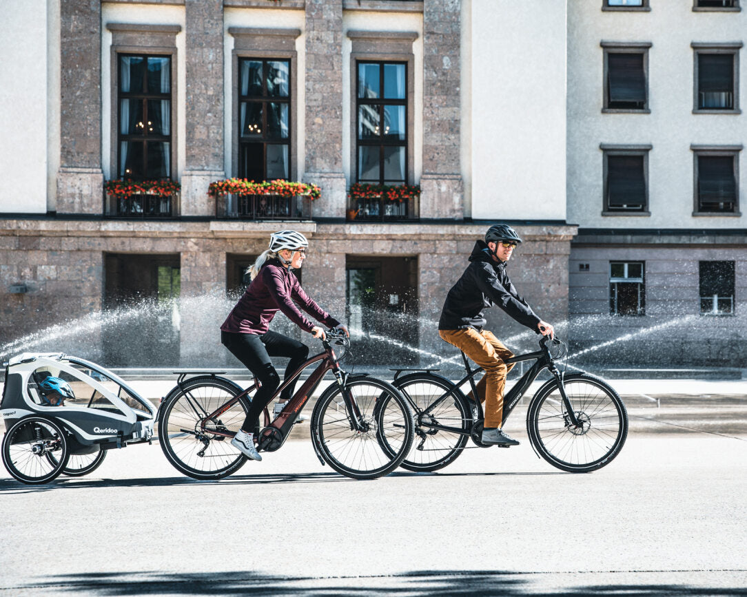 Zwei Personen fahren auf Specialized E-Bikes mit Kind im Qeridoo-Anhänger an einem Springbrunnen vorbei.