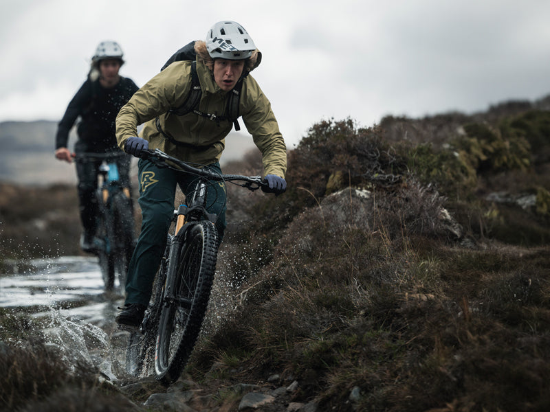 Zwei Mountainbiker fahren durch feuchtes, hügeliges Gelände; wetterfeste Kleidung, Helme, energische Haltung.