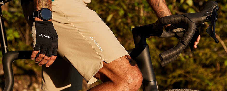 Mann in Vaude-Radshorts hält Fahrradlenker, trägt Radhandschuhe und Uhr; Outdoor-Umgebung.