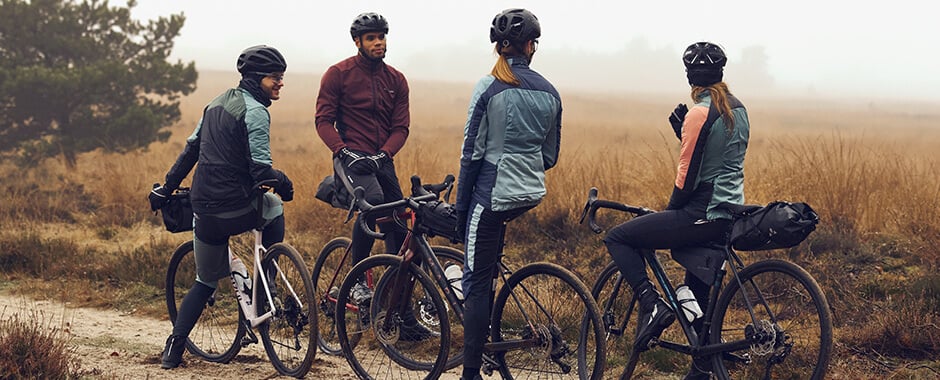 Gruppe von Radfahrern in Outdoor-Bekleidung auf Schotterweg, Vaude-Kleidung erkennbar, bei nebligem Wetter im ländlichen Gebiet.