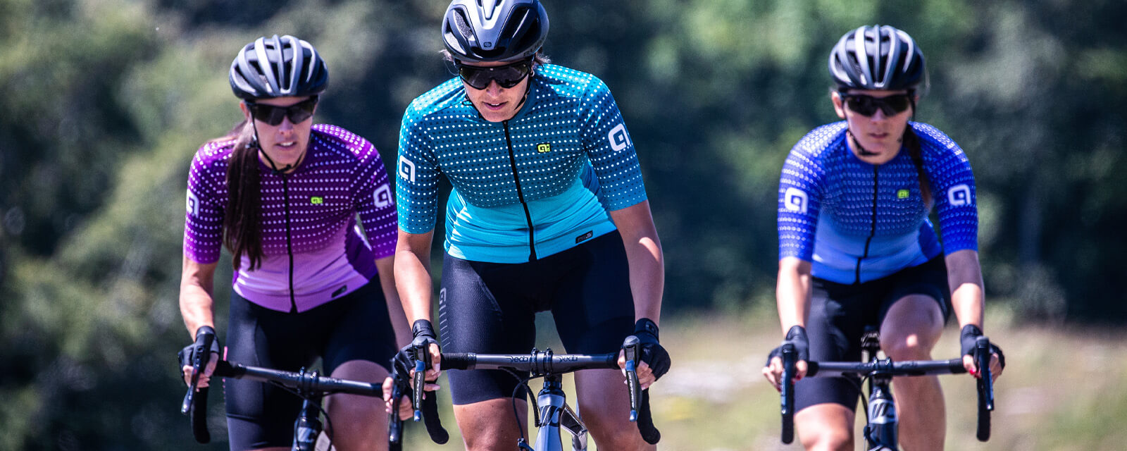Drei Radfahrerinnen in bunten Ale-Trikots und Helmen beim Training auf Rennrädern.