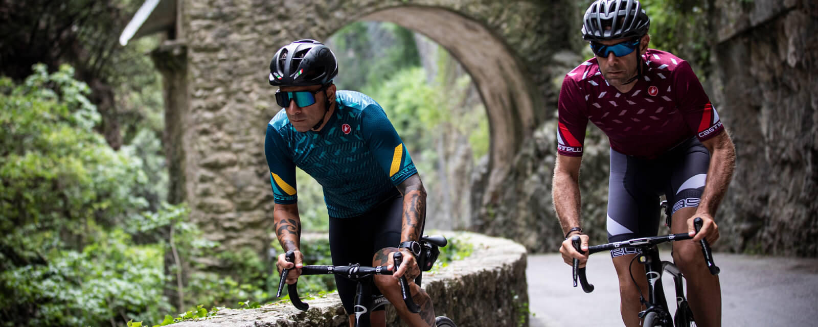 Zwei Radfahrer in Castelli-Radtrikots fahren durch eine malerische Steinstraße im Grünen.
