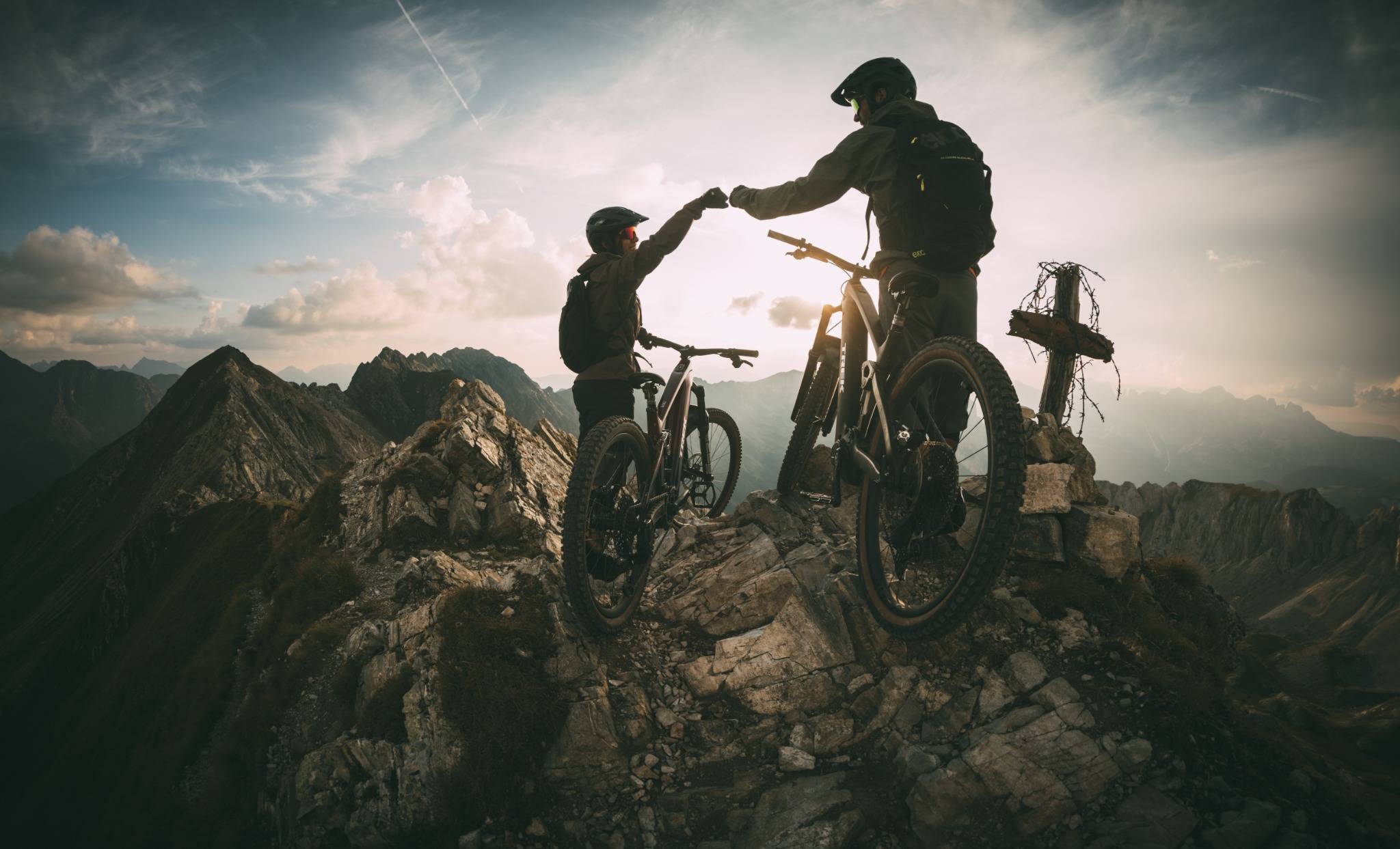 Zwei Mountainbiker auf Berggipfel, Fäuststoß, Sonnenuntergang, Trek Rail E-Bikes, Abenteuer in alpiner Landschaft.