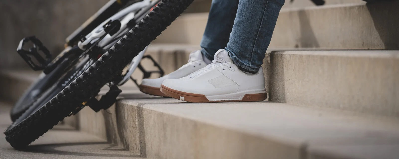 Weiße Crankbrothers-Schuhe auf einer Treppe, neben einem Fahrrad mit breiten Reifen, ideal für Radfahrer.