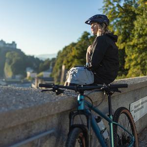 Frau mit Helm sitzt auf einer Mauer, neben ihr ein Cube-Fahrrad. Im Hintergrund Bäume und Gebäude in urbaner Umgebung.