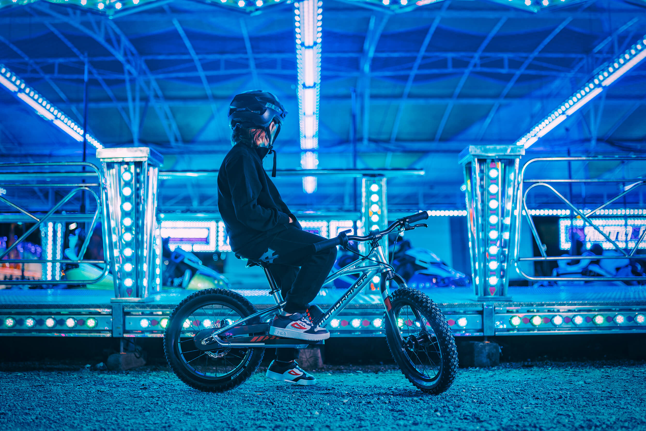 Kind auf Mondraker Kinder-E-Bike im beleuchteten Freizeitpark, dunkle Kleidung, Fahrrad für junge Abenteurer.