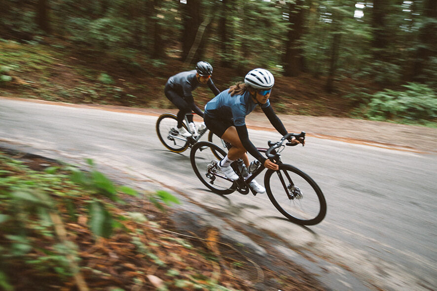 Zwei Rennradfahrer in Aktion auf einer kurvigen Waldstraße, tragen Helme und Sportbekleidung.