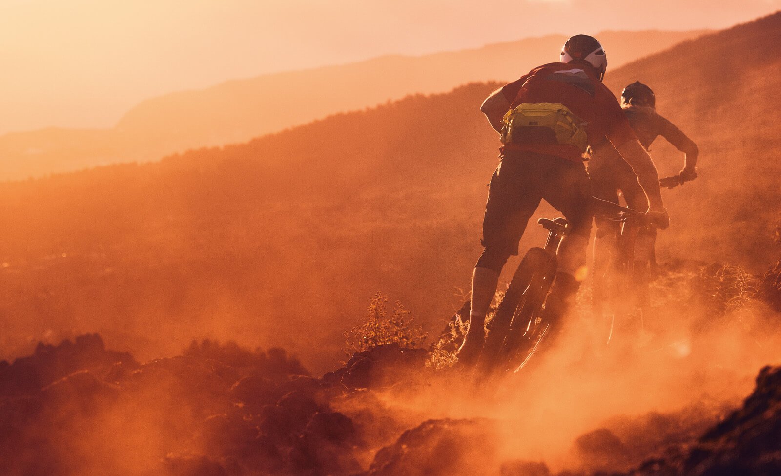 Zwei Mountainbiker fahren im Sonnenuntergang, tragen Helme und Rucksäcke, bergab durch staubiges Gelände.