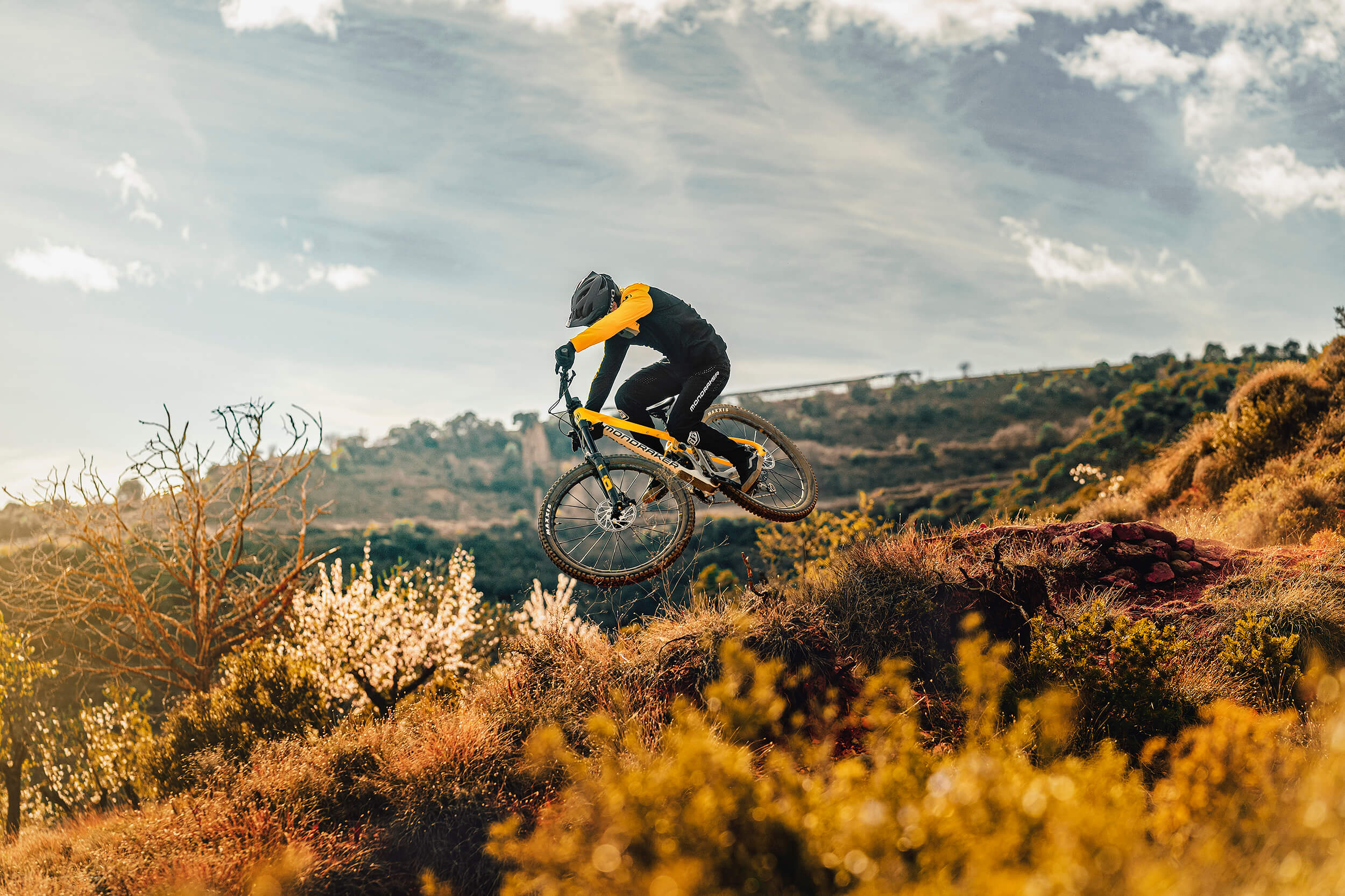 Mountainbiker auf einem gelben Mondraker-Mountainbike springt durch hügelige Landschaft bei sonnigem Wetter.