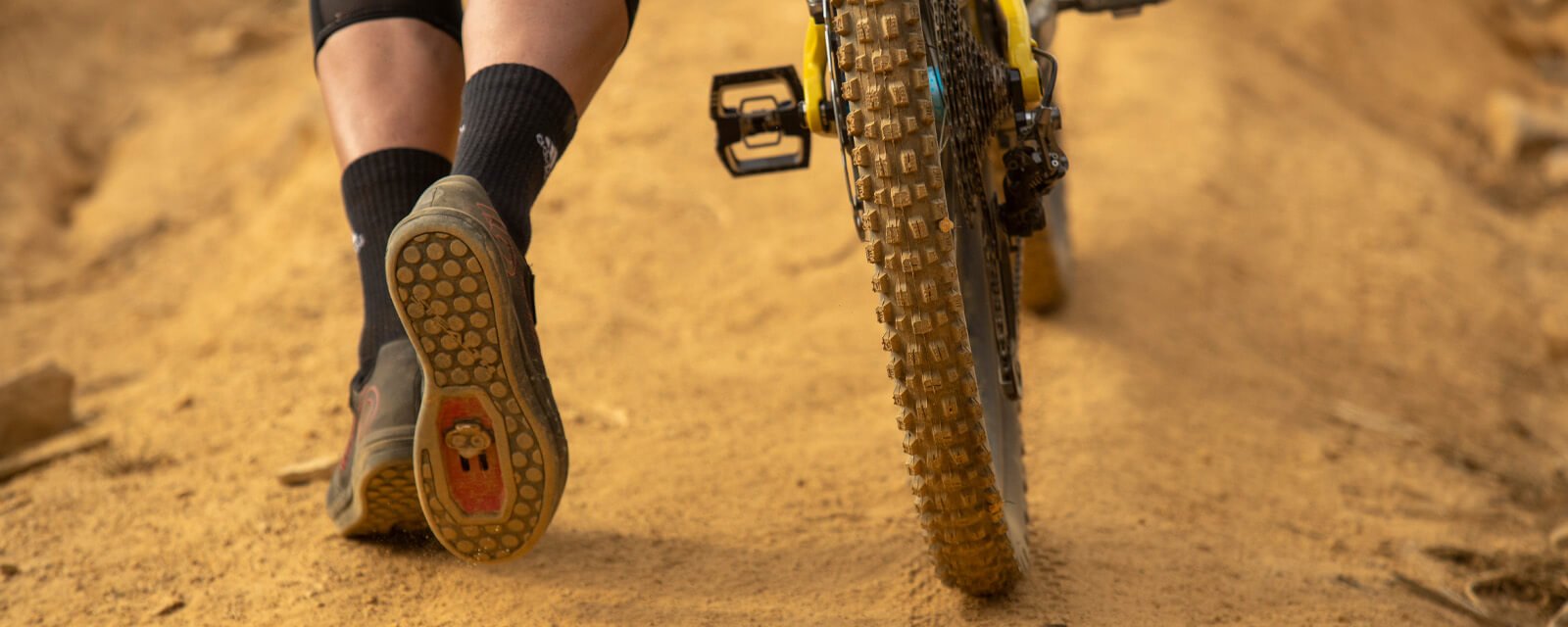 Mountainbike auf sandigem Weg, Person trägt Five Ten Fahrradschuhe, Detailaufnahme Reifen und Schuhprofil.