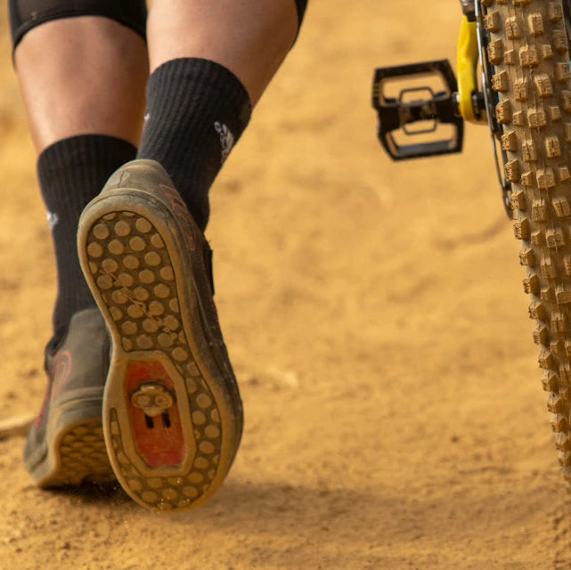 Biker in Klickpedalen, Mountainbike-Schuh mit SPD-Cleats auf sandigem Weg, gelber Fahrradrahmen sichtbar.