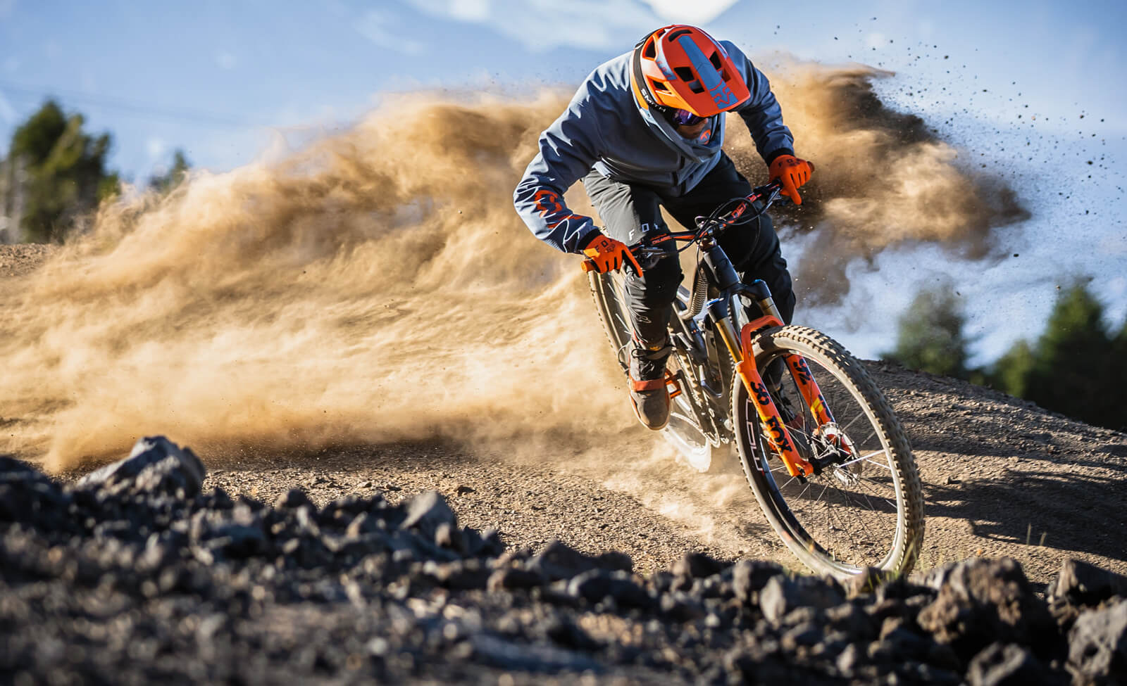Ein Mountainbiker in orangener Schutzkleidung fährt mit hohem Tempo durch eine staubige Kurve.