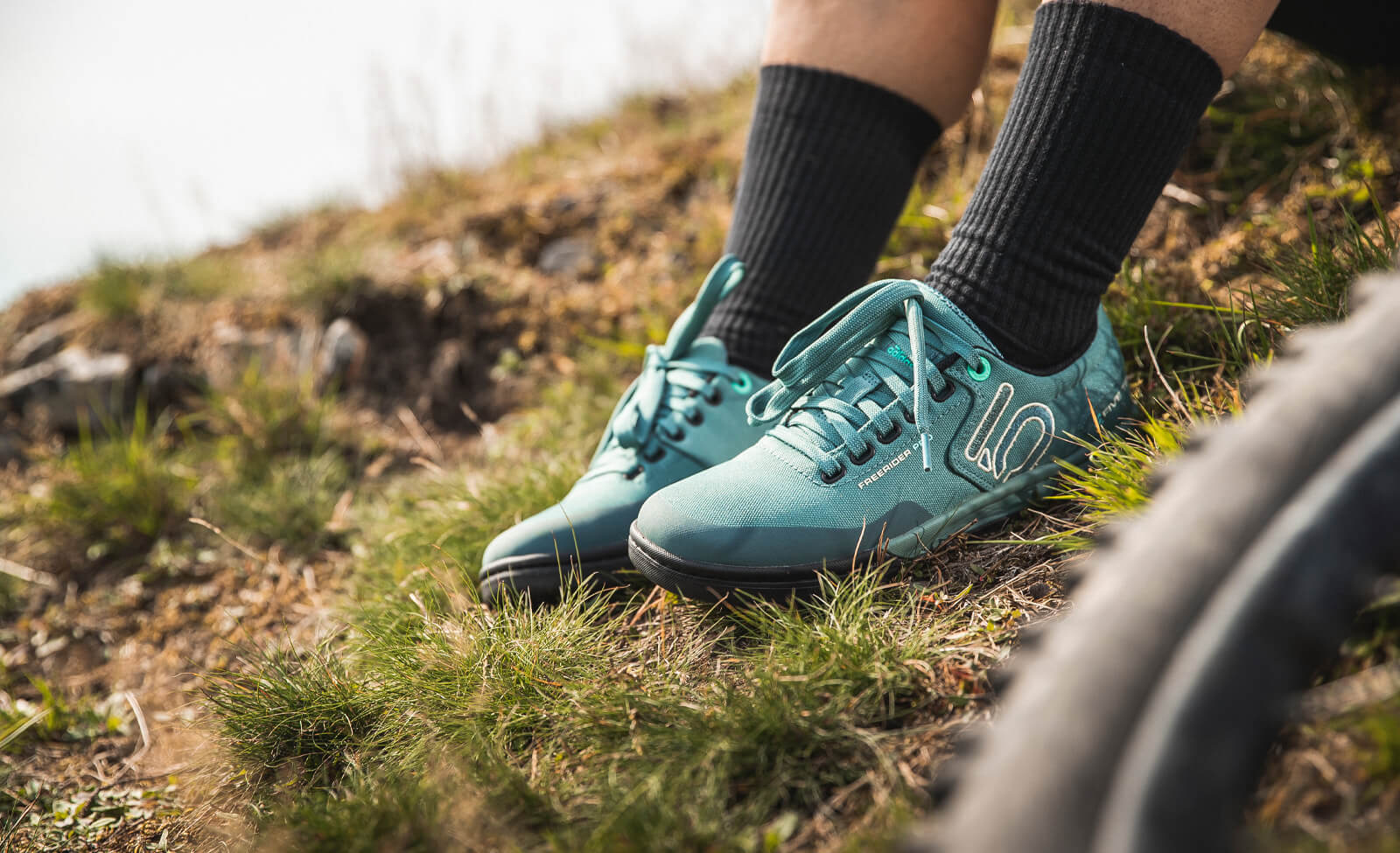 Mountainbiker trägt türkisfarbene Freerider-Schuhe von Five Ten auf einem grasbewachsenen Hügel.