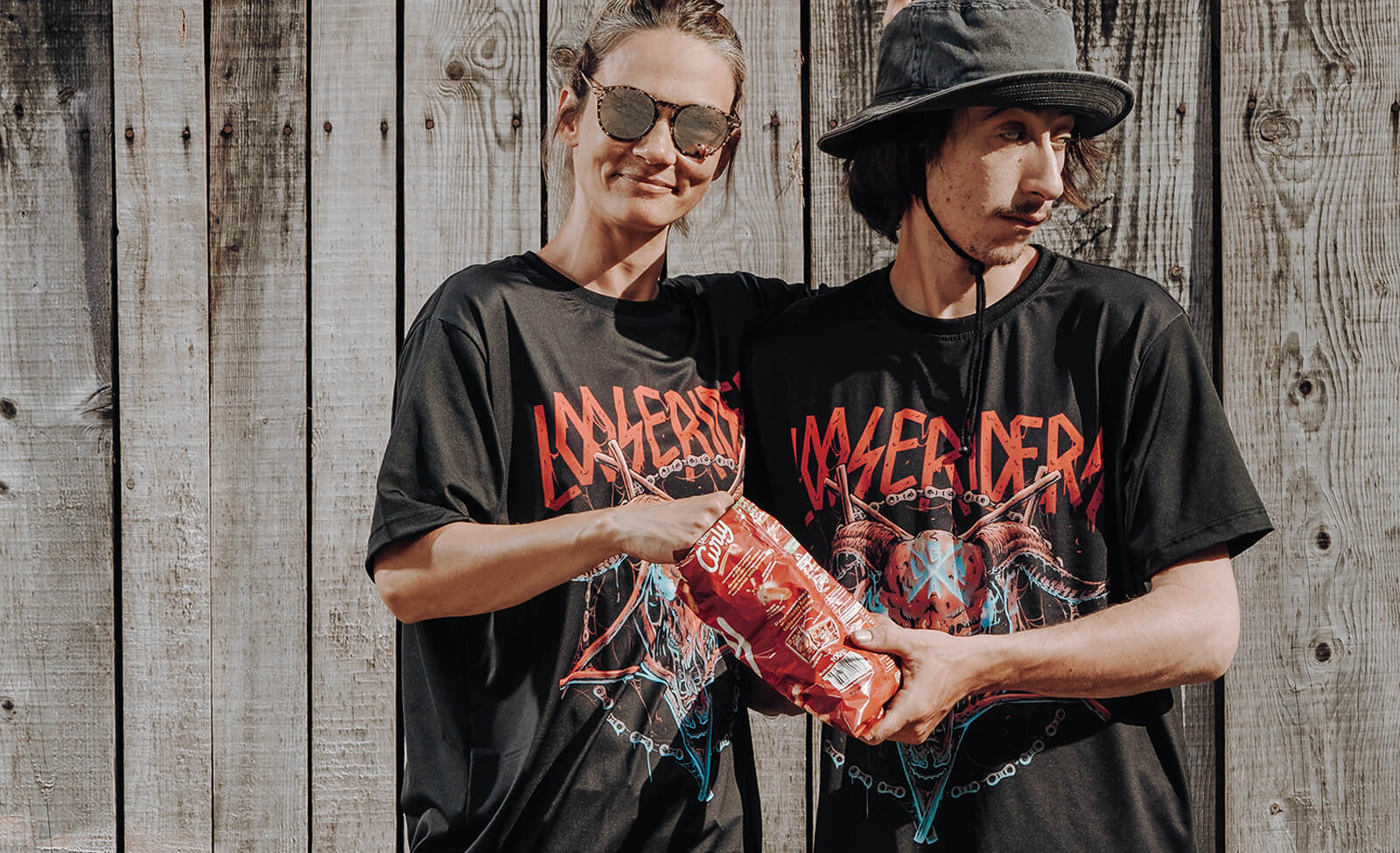 Zwei Personen in schwarzen "LOSERDERA"-T-Shirts teilen eine Snacktüte vor einer Holzwand.