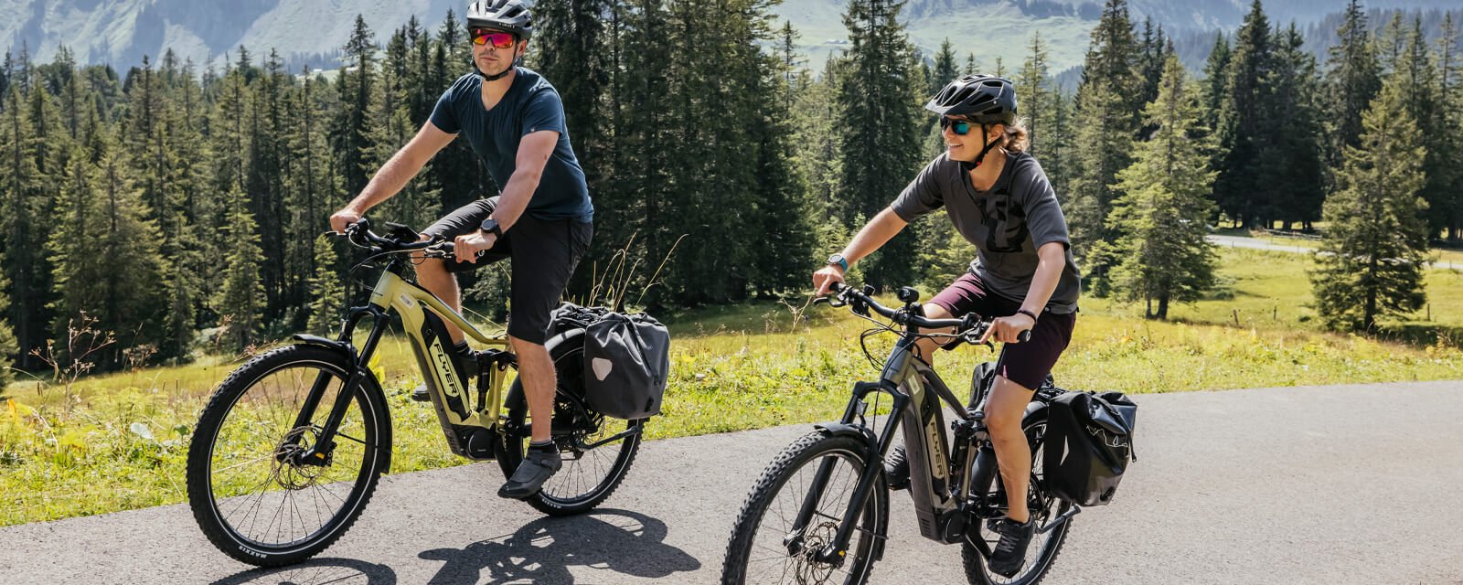 Zwei Personen fahren Flyer E-Bikes auf einer Straße in einer bergigen, bewaldeten Landschaft.