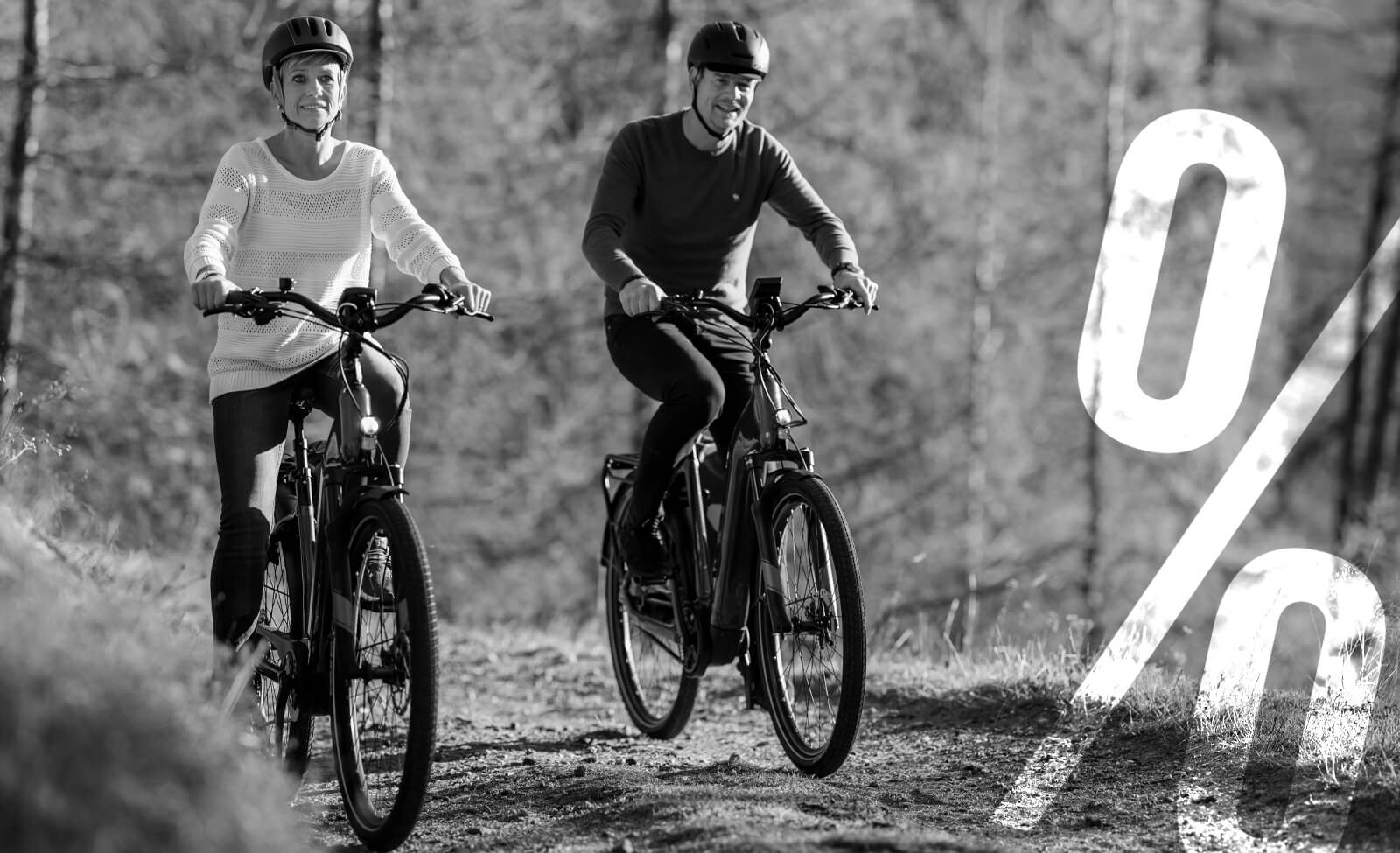 Zwei Personen fahren mit E-Bikes im Wald; Fahrradhelme tragend. Prozentzeichen-Dekor im Vordergrund.