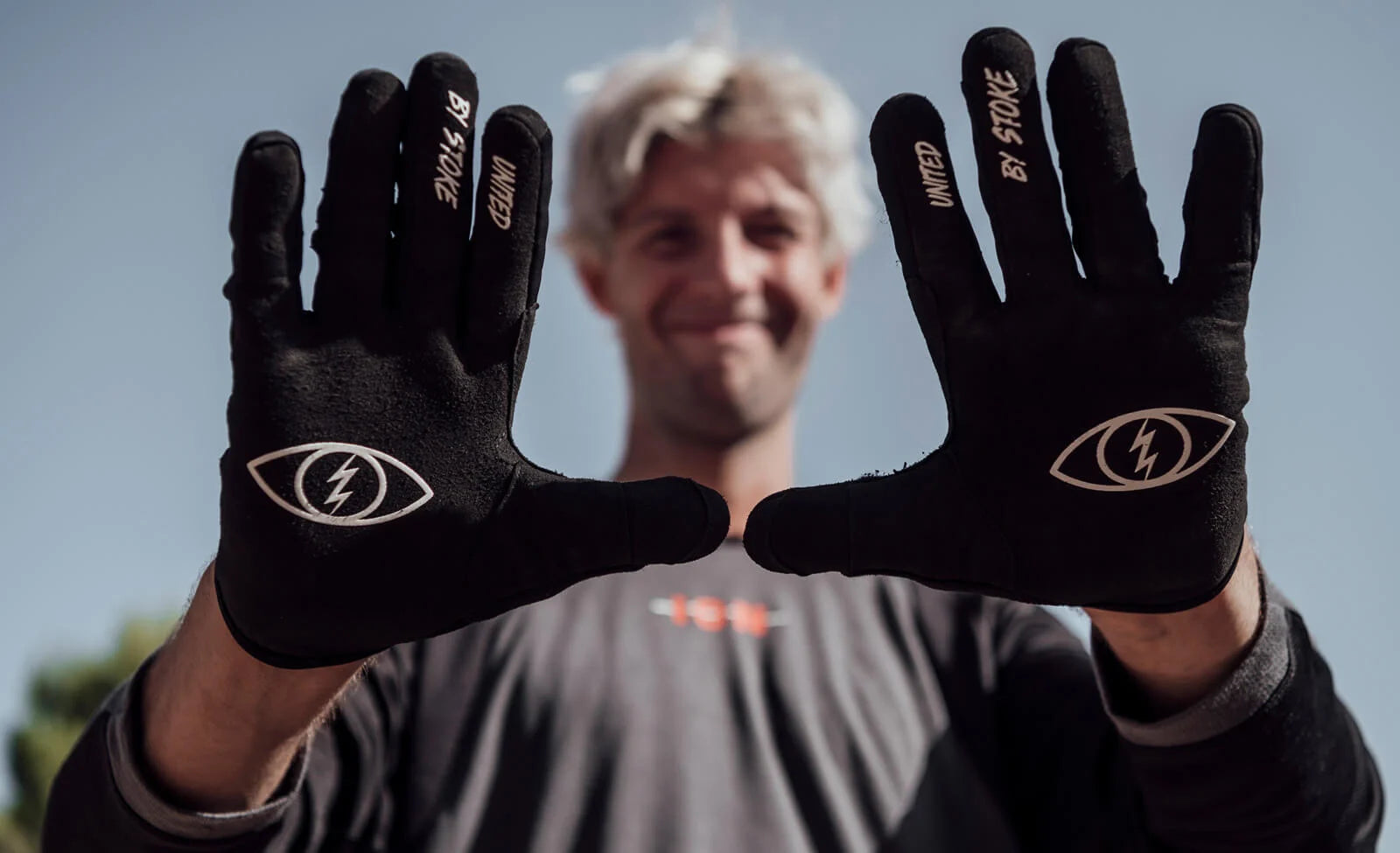 Mann zeigt Handschuhe mit Blitz-Augensymbol und Schriftzug "United by Stoke" vor blauem Himmel.