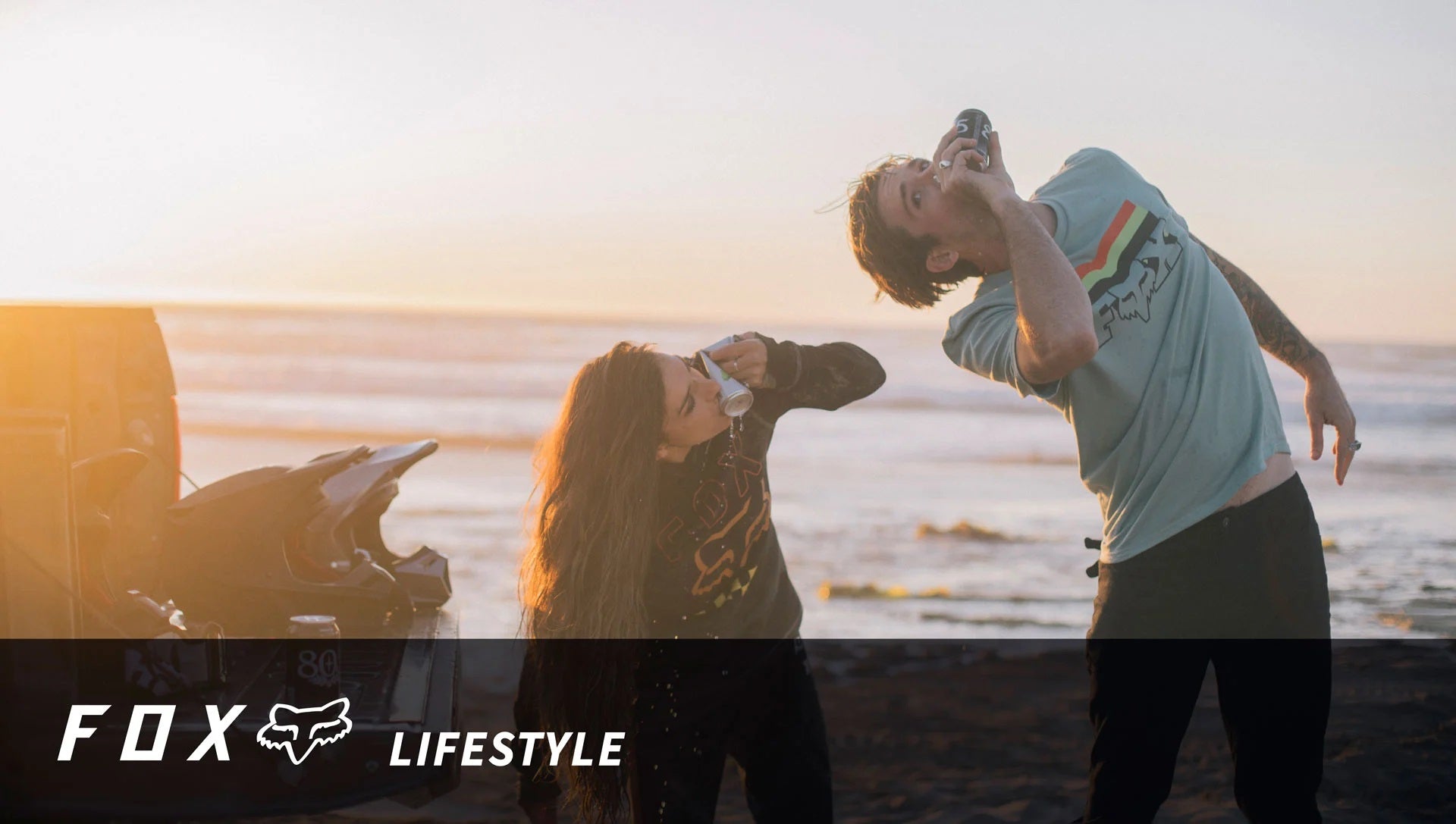 Zwei Personen in Fox-Kleidung am Strand trinken aus Dosen, bei Sonnenuntergang.