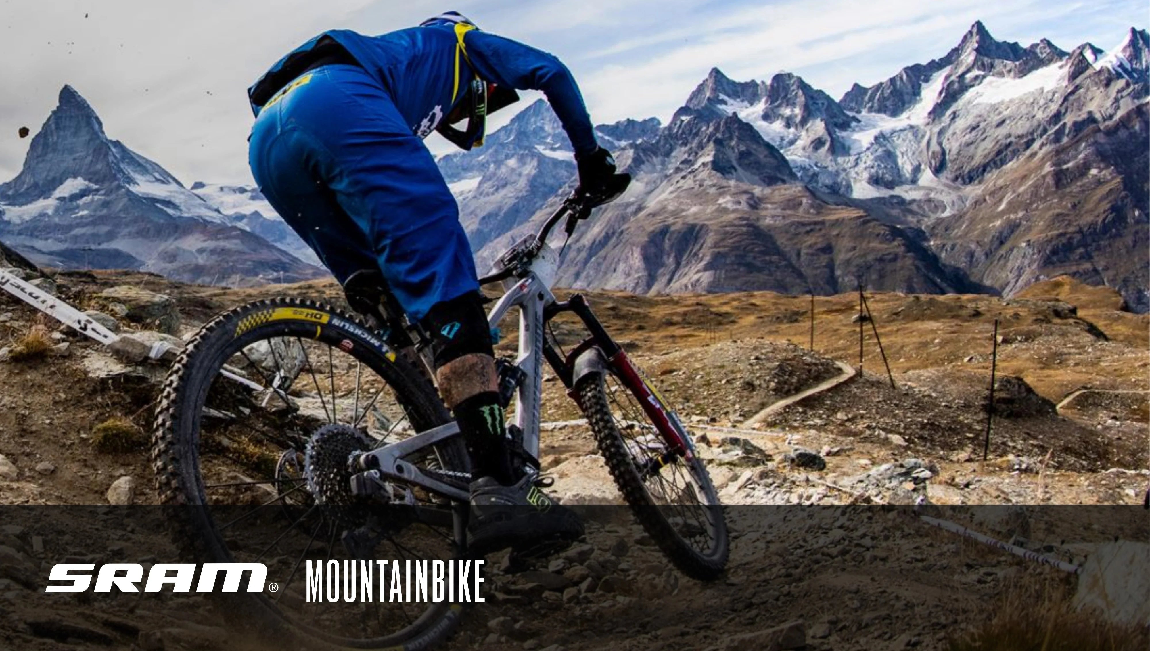 Mountainbiker in blauer Ausrüstung fährt SRAM-Rad auf steinigem Gebirgspfad mit spektakulärer Berglandschaft im Hintergrund.