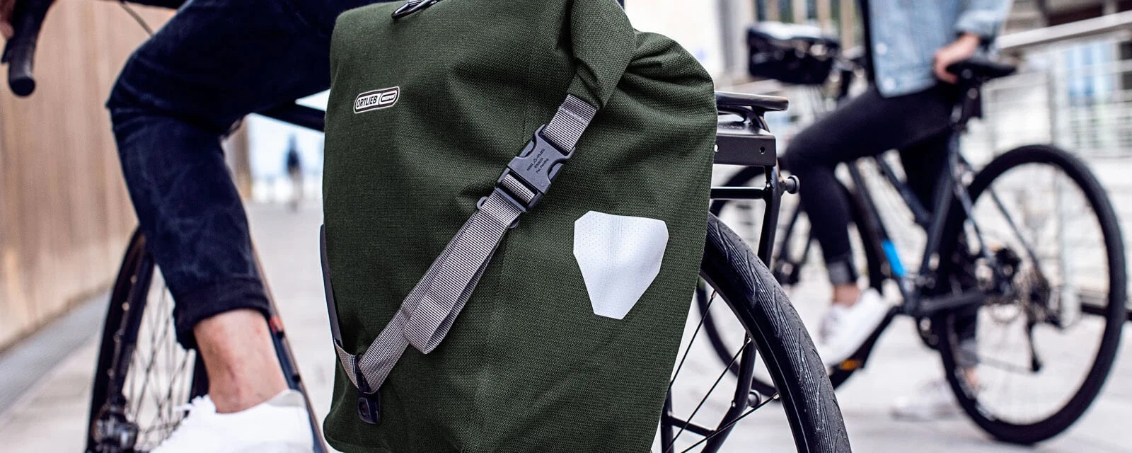 Grüne Ortlieb-Fahrradtasche am Gepäckträger montiert, ideal für Pendler. Zwei Personen mit Fahrrädern im Hintergrund.