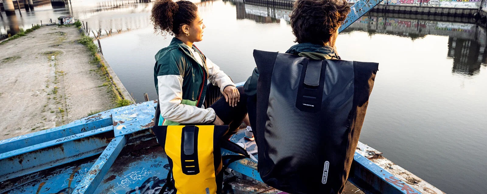 Zwei Personen mit Ortlieb-Rucksäcken sitzen auf einer Brücke am Wasser, genießen die Aussicht.