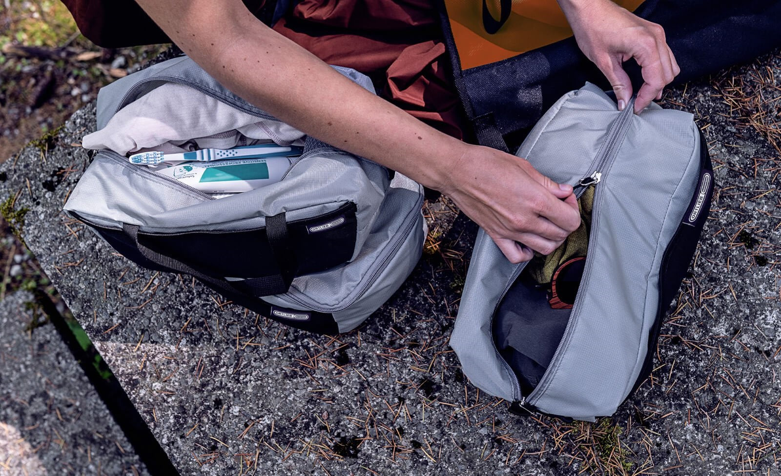 Zwei graue Ortlieb-Packtaschen mit Reißverschlüssen, ideal zum Sortieren von Campingausrüstung, auf einer Steinoberfläche.