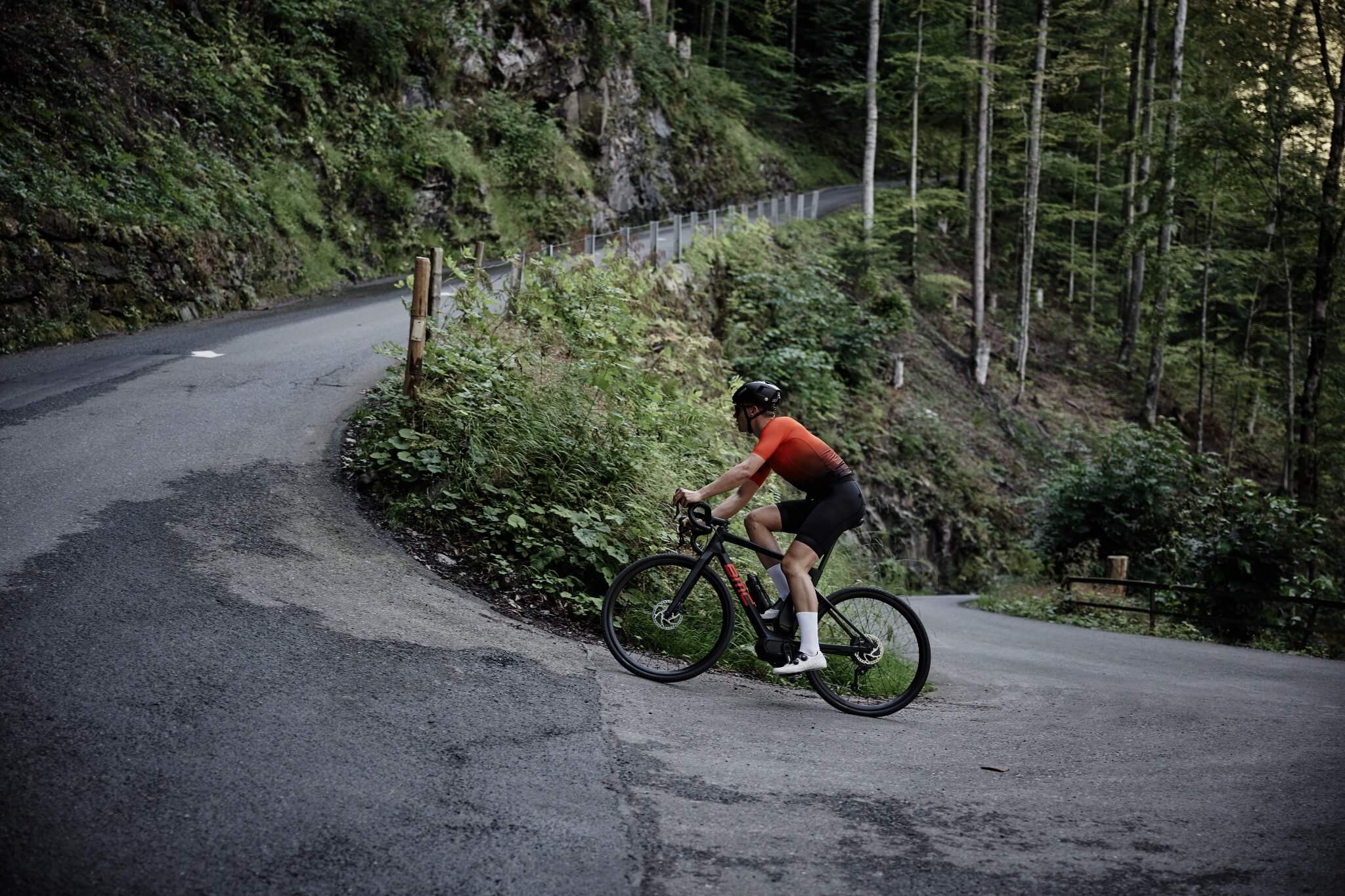 Radfahrer fährt mit BMC Alpenchallenge AMP auf einer kurvigen Waldstraße bergauf, ideal für sportliche Radfahrer.